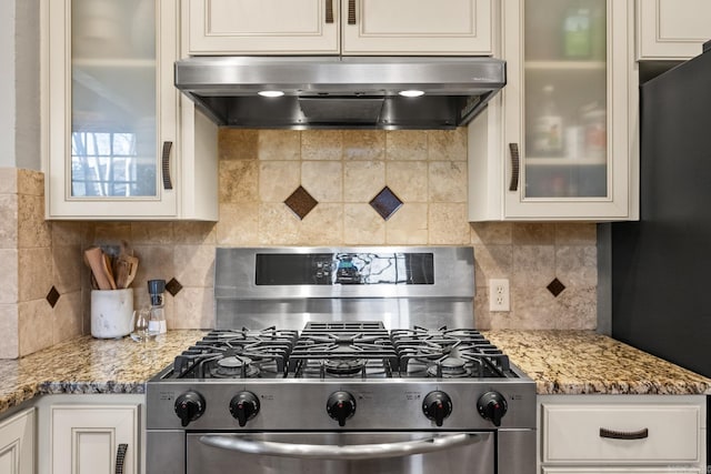 kitchen with stainless steel gas stove, light stone countertops, range hood, and glass insert cabinets