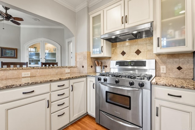 kitchen featuring arched walkways, ornamental molding, light stone countertops, under cabinet range hood, and stainless steel range with gas stovetop