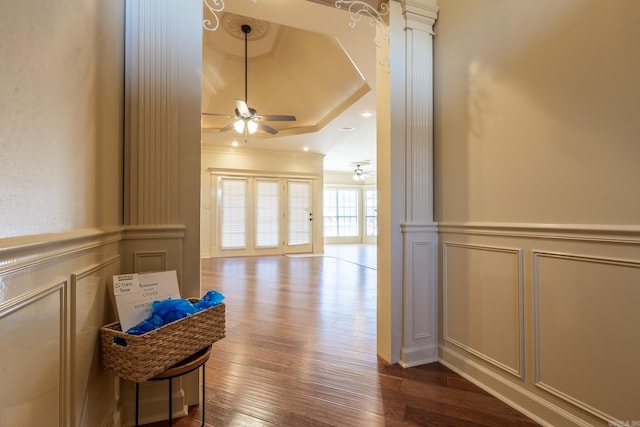 hall with a tray ceiling, crown molding, a decorative wall, dark wood-type flooring, and ornate columns