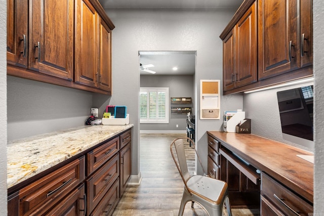 kitchen featuring built in desk, wood finished floors, a ceiling fan, light stone countertops, and baseboards