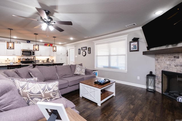 living room featuring crown molding, visible vents, dark wood-type flooring, a stone fireplace, and baseboards