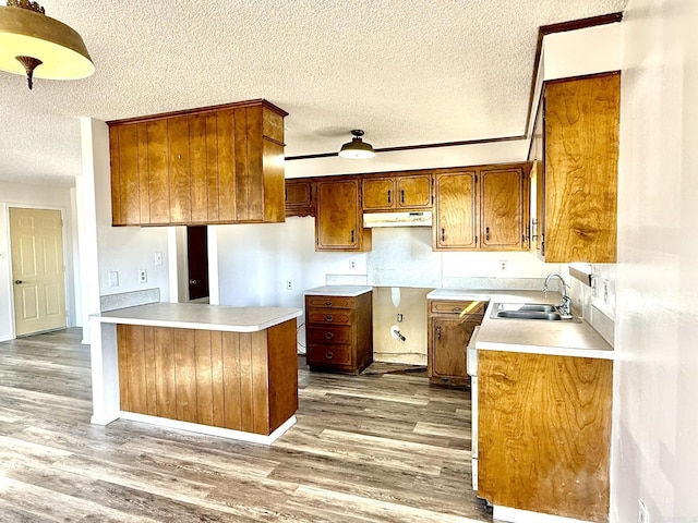 kitchen featuring brown cabinetry, wood finished floors, a textured ceiling, under cabinet range hood, and a sink