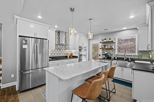 kitchen featuring wall chimney exhaust hood, a center island, stainless steel appliances, white cabinetry, and a sink