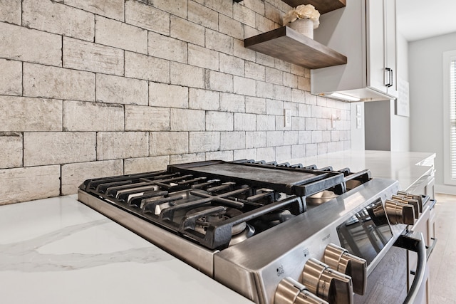 kitchen featuring decorative backsplash, light stone counters, wall chimney range hood, open shelves, and stainless steel range with gas stovetop