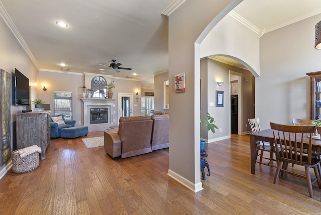 living area with arched walkways, a ceiling fan, a glass covered fireplace, baseboards, and hardwood / wood-style flooring