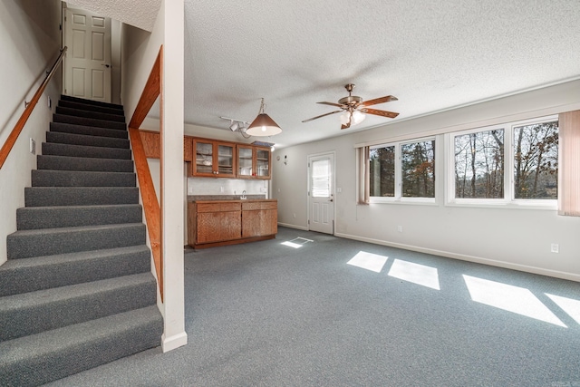 unfurnished living room featuring a textured ceiling, ceiling fan, baseboards, stairs, and dark colored carpet