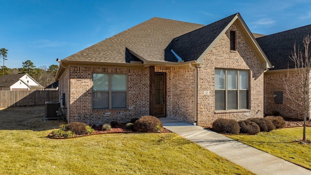 view of front of property featuring brick siding, roof with shingles, central air condition unit, fence, and a front lawn