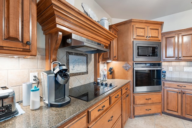 kitchen featuring appliances with stainless steel finishes, dark stone countertops, brown cabinets, and under cabinet range hood