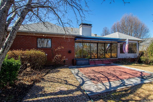 rear view of property featuring a sunroom, roof with shingles, a chimney, and brick siding