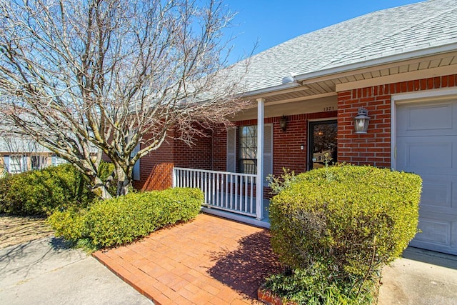 view of exterior entry featuring a garage, brick siding, a porch, and roof with shingles