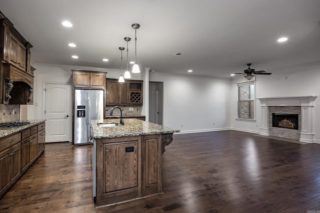 kitchen featuring light stone counters, a sink, a lit fireplace, stainless steel refrigerator with ice dispenser, and an island with sink