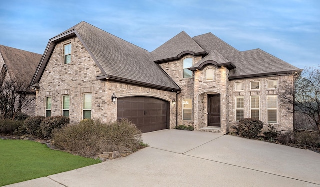 french country inspired facade featuring a shingled roof, concrete driveway, brick siding, and an attached garage