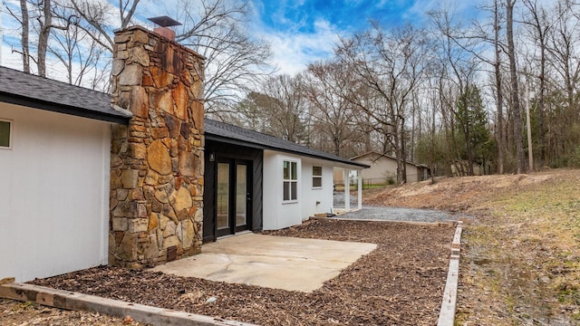 exterior space with stone siding, a shingled roof, a chimney, and a patio