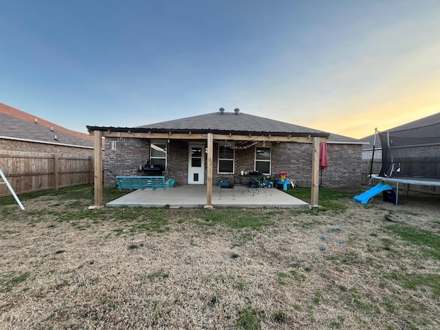 back of house at dusk with a fenced backyard, brick siding, a yard, a trampoline, and a patio area