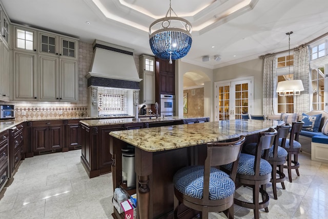 kitchen featuring a center island with sink, a raised ceiling, decorative backsplash, french doors, and premium range hood