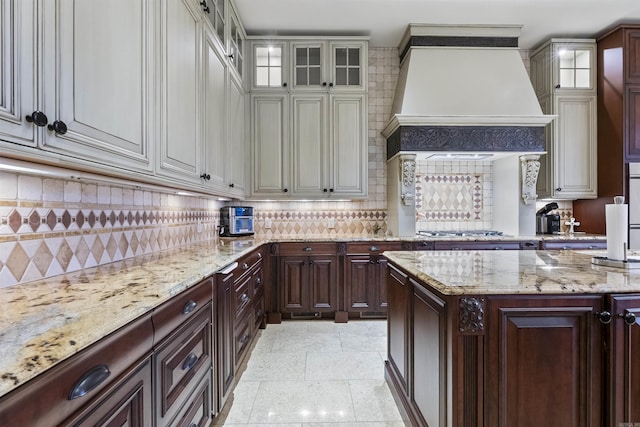 kitchen featuring custom range hood, light stone countertops, tasteful backsplash, glass insert cabinets, and stainless steel gas stovetop