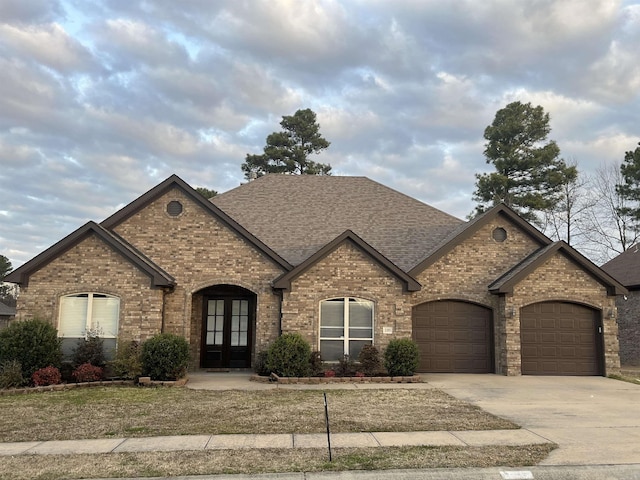 french country inspired facade featuring driveway, a shingled roof, an attached garage, french doors, and brick siding