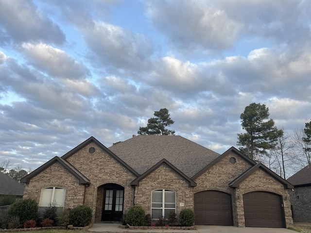 french country home featuring driveway, a shingled roof, an attached garage, french doors, and brick siding