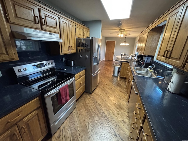 kitchen with light wood finished floors, dark countertops, appliances with stainless steel finishes, under cabinet range hood, and a sink