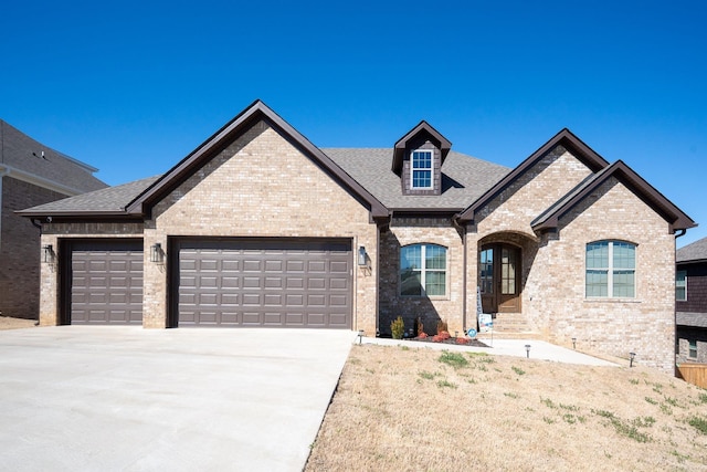 french country home with a garage, roof with shingles, concrete driveway, and brick siding