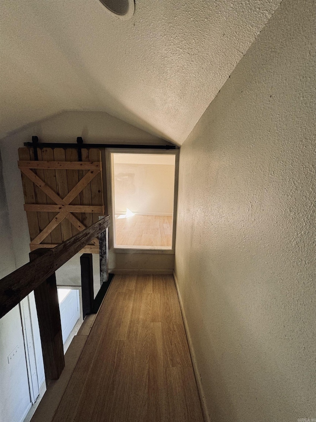 hallway featuring a textured ceiling, a textured wall, wood finished floors, baseboards, and vaulted ceiling