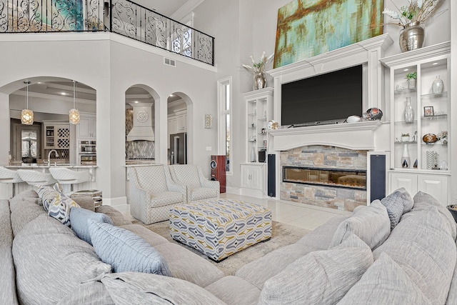 living room with arched walkways, a stone fireplace, visible vents, baseboards, and crown molding