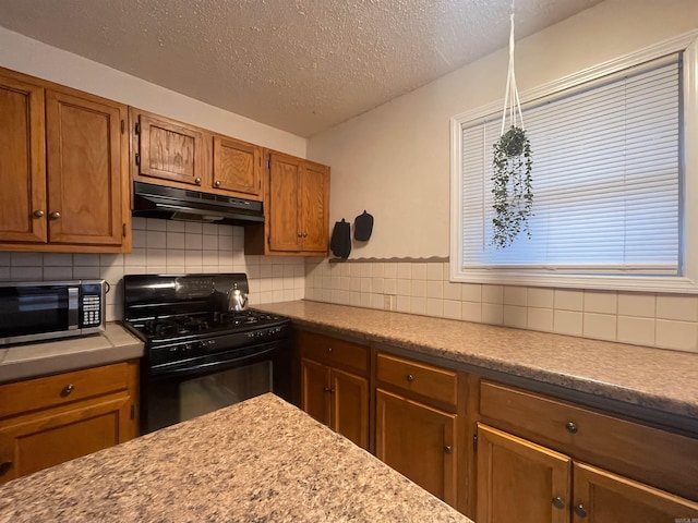 kitchen featuring under cabinet range hood, brown cabinetry, decorative backsplash, stainless steel microwave, and gas stove