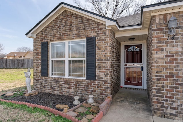 doorway to property featuring a shingled roof, brick siding, and fence