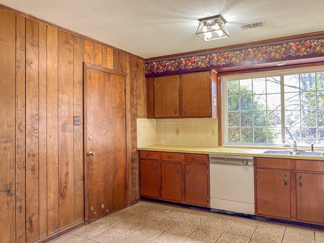 kitchen with light countertops, brown cabinetry, white dishwasher, wooden walls, and a sink