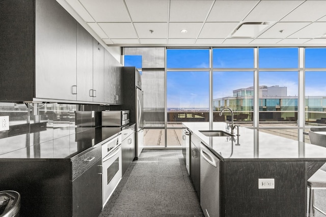 kitchen with floor to ceiling windows, a center island with sink, appliances with stainless steel finishes, a sink, and dark cabinetry