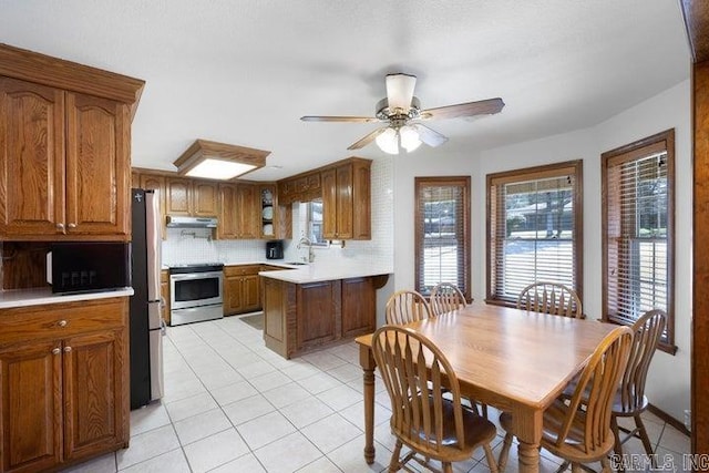 kitchen with light tile patterned floors, under cabinet range hood, stainless steel appliances, a peninsula, and brown cabinets