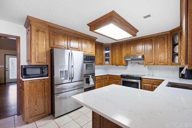 kitchen featuring under cabinet range hood, stainless steel appliances, backsplash, brown cabinets, and glass insert cabinets