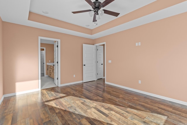 unfurnished bedroom with connected bathroom, a ceiling fan, baseboards, a tray ceiling, and wood-type flooring