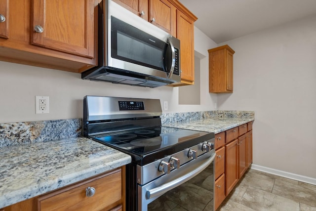 kitchen with appliances with stainless steel finishes, brown cabinetry, light stone counters, and baseboards