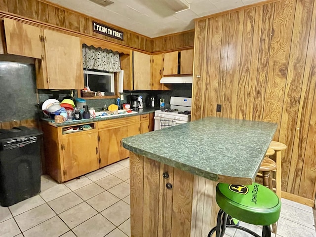 kitchen with light tile patterned floors, under cabinet range hood, a breakfast bar, a center island, and white gas range