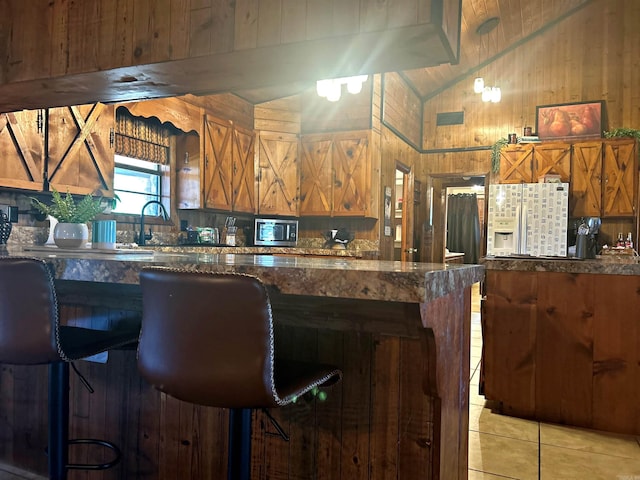 kitchen featuring light tile patterned floors, wooden walls, refrigerator with ice dispenser, vaulted ceiling, and stainless steel microwave