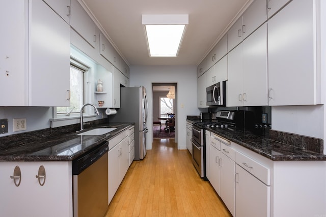 kitchen featuring light wood finished floors, white cabinets, dark stone counters, stainless steel appliances, and a sink