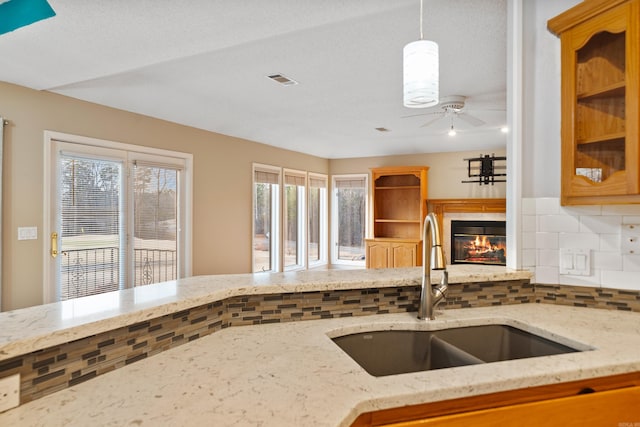 kitchen featuring tasteful backsplash, a glass covered fireplace, ceiling fan, light stone counters, and a sink