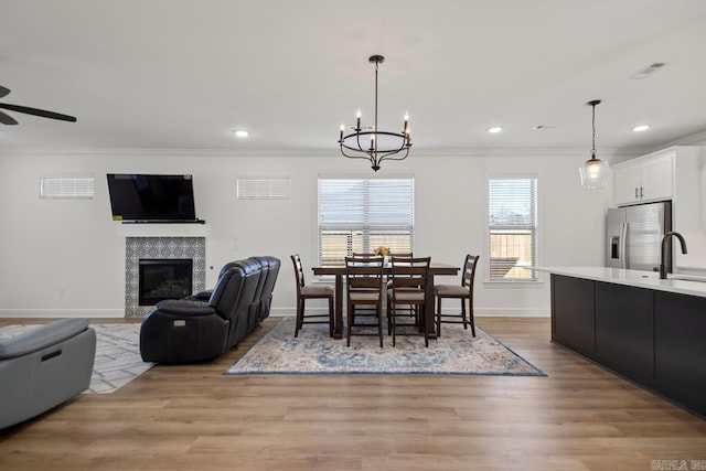 dining space featuring baseboards, a tile fireplace, crown molding, light wood-style floors, and ceiling fan with notable chandelier