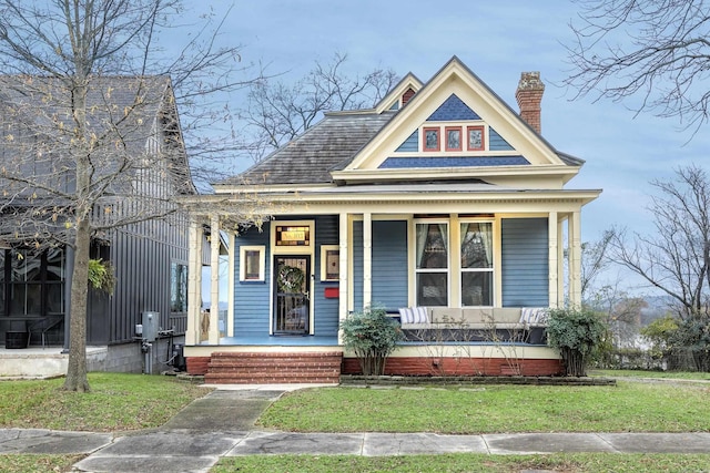 view of front facade featuring covered porch, roof with shingles, a chimney, and a front yard