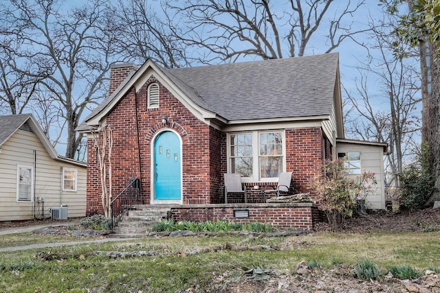 view of front facade featuring a shingled roof, brick siding, a chimney, and central AC unit