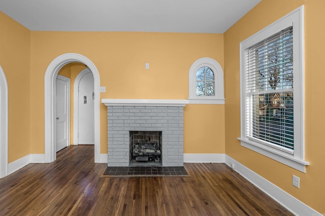 unfurnished living room featuring arched walkways, a brick fireplace, wood finished floors, and baseboards