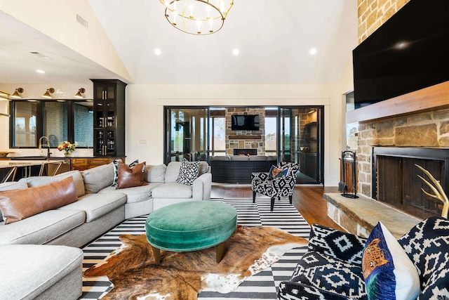 living area featuring high vaulted ceiling, a stone fireplace, wood finished floors, visible vents, and an inviting chandelier