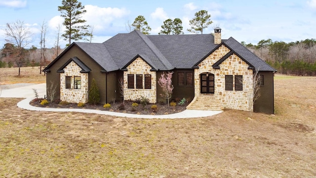 french country inspired facade featuring stone siding, a shingled roof, a chimney, and a front lawn