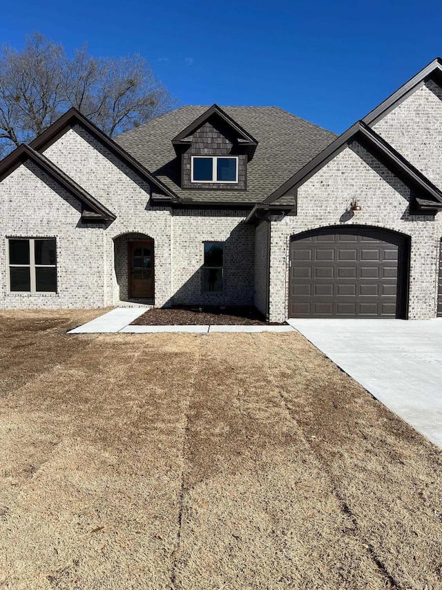 french country style house featuring driveway, a shingled roof, an attached garage, and brick siding