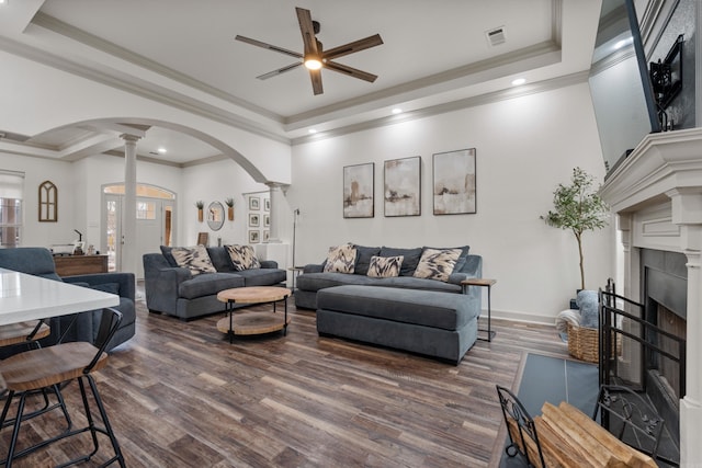 living room with arched walkways, dark wood-style flooring, decorative columns, a raised ceiling, and a fireplace with flush hearth