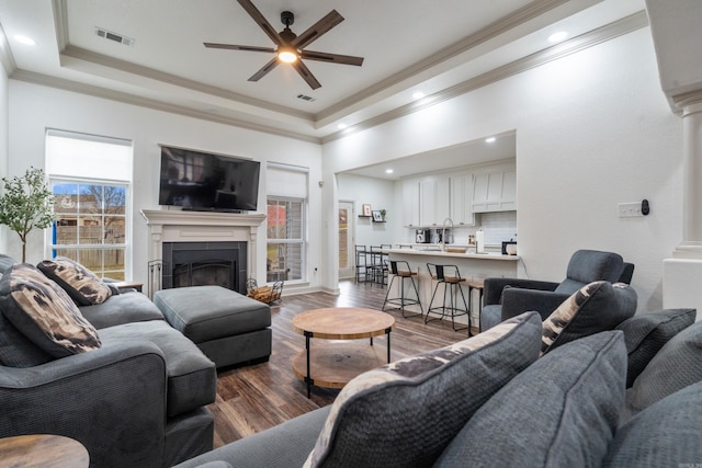 living room featuring a tray ceiling, crown molding, a fireplace, dark wood finished floors, and decorative columns