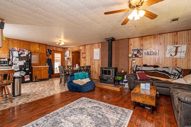 living area featuring visible vents, a wood stove, ceiling fan, wood walls, and hardwood / wood-style flooring