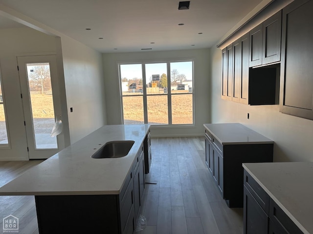 kitchen with a sink, baseboards, light wood-style floors, light countertops, and an island with sink