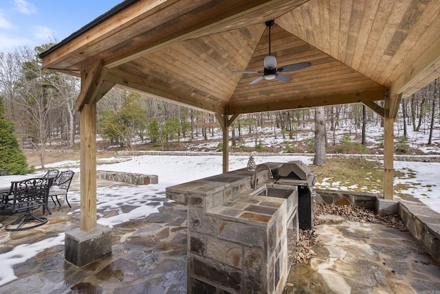 snow covered patio with an outdoor kitchen, a ceiling fan, a gazebo, grilling area, and outdoor dining space